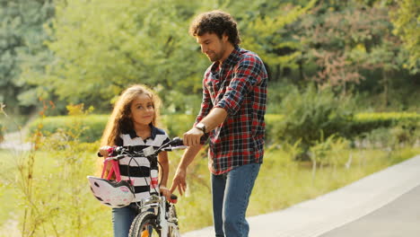 Portrait-of-a-little-girl-and-her-dad-standing-near-the-bike.-They-look-at-each-other-and-then---into-the-camera-and-smile.-Blurred-background